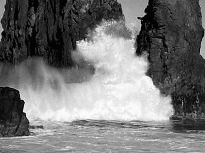 Schwarz-weiß-Foto einer Welle, die zwischen schroffen Felsen hindurchbricht und Gischt aufwirbelt