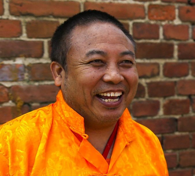 Porträt von Rinpoche, lachend, in traditionellem safranfarbenem Gewand vor einer roten Backsteinmauer