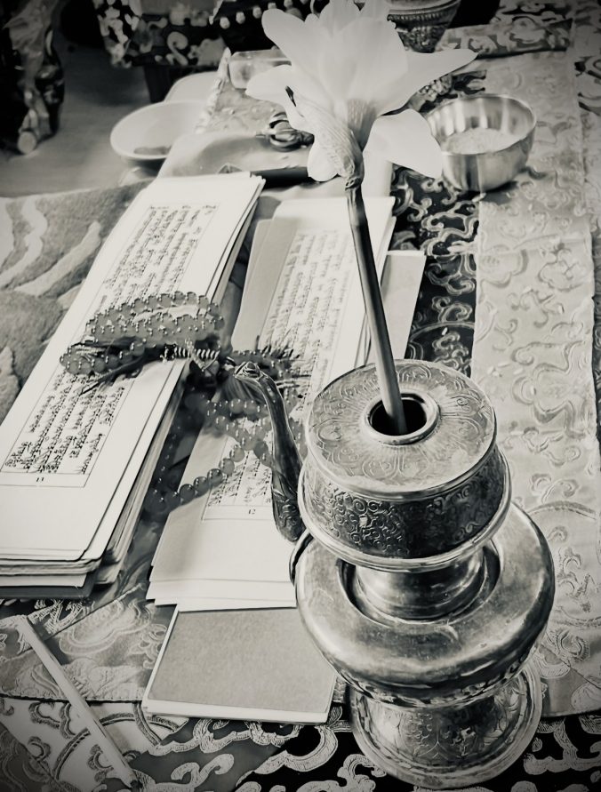 Buddhistischer Schreintisch mit aufgeschlagenen tibetischen Texten, Mala und Ritualvase auf Brokatstoff schwarz-weiß